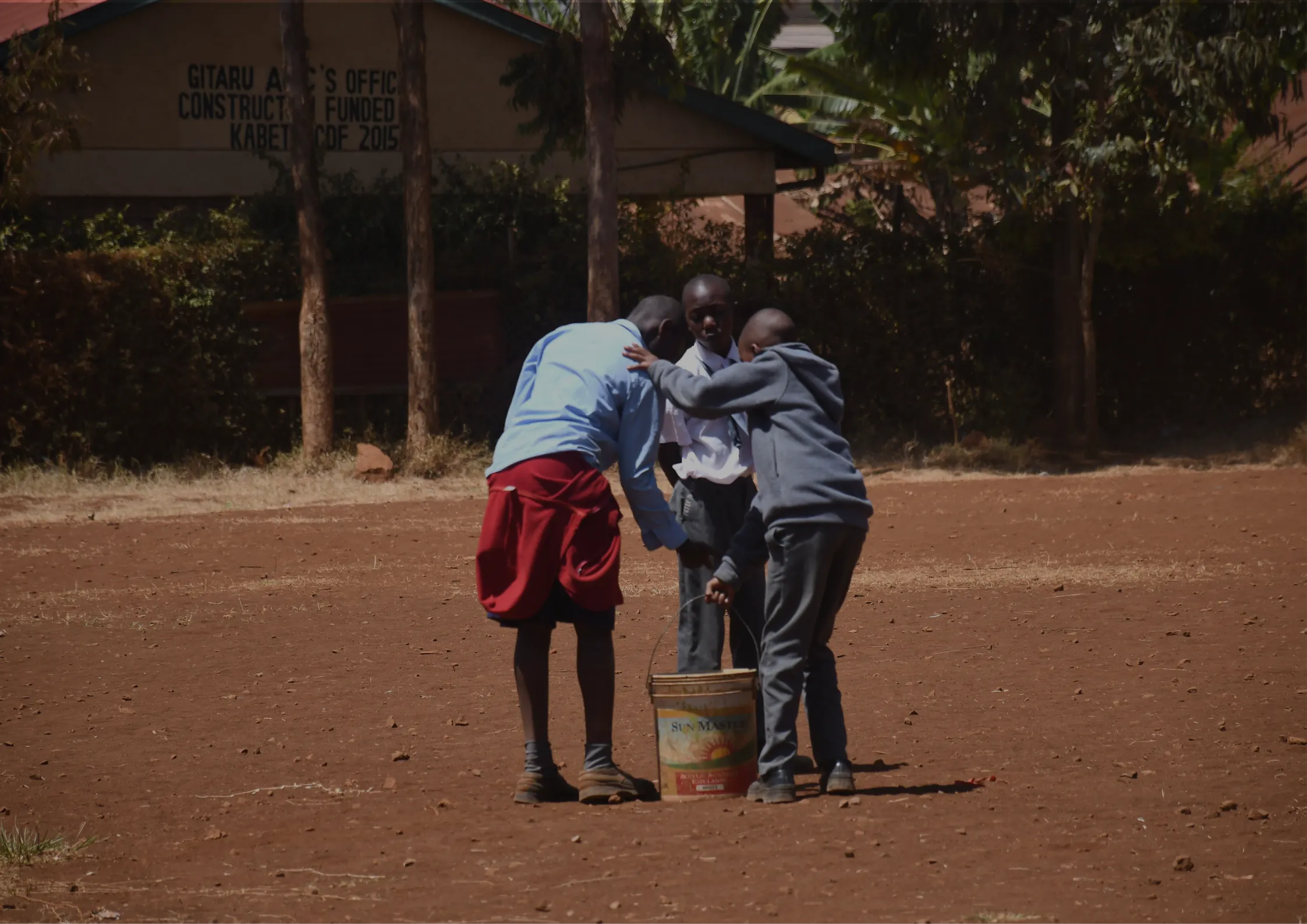 Three boys stand closely together on a dirt field, gathered around a yellow drum. Trees and a building with a sign are visible in the background. The boys appear to be focused on the drum. -Kaki Foundation