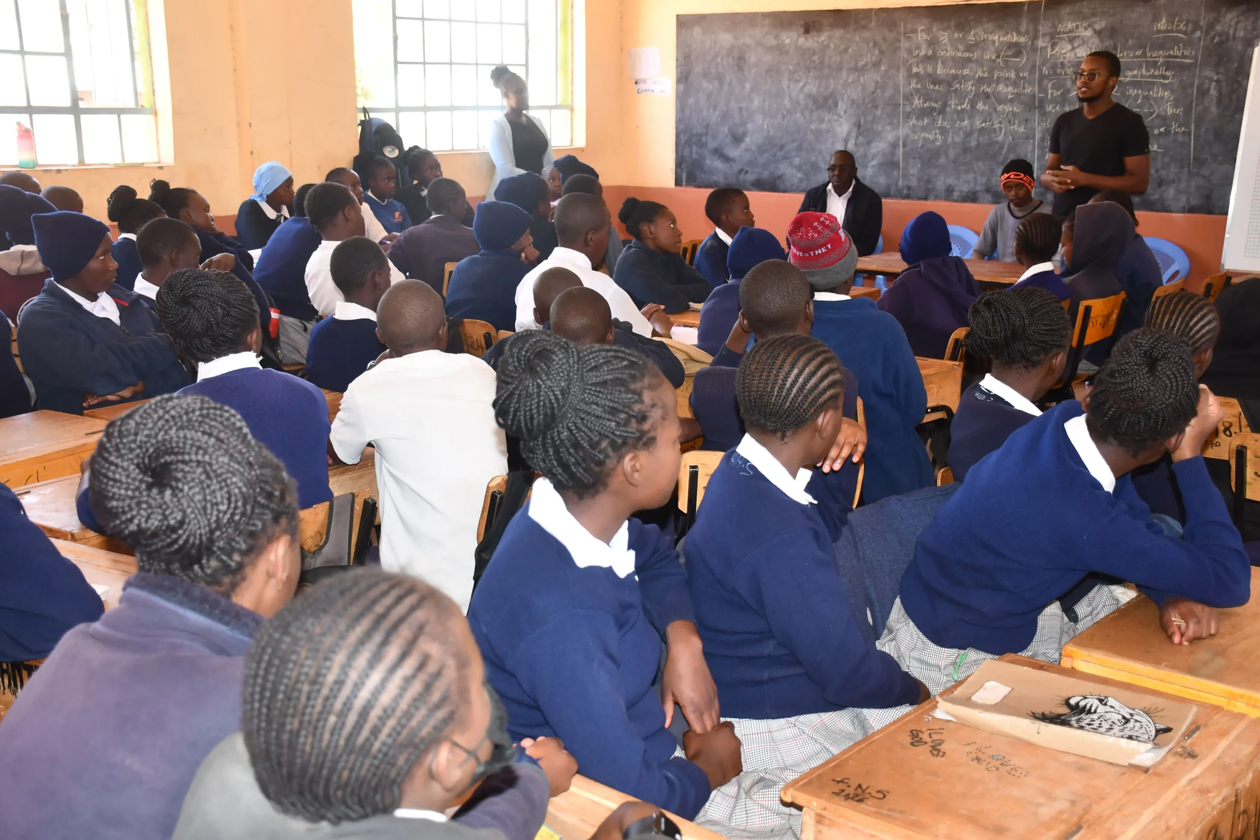 A classroom full of students in blue uniforms attentively listening to a speaker at the front, with several adults seated near a chalkboard and windows letting in natural light. -Kaki Foundation