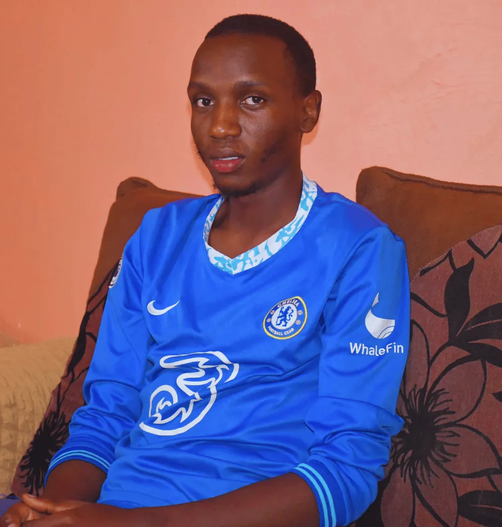 A young man wearing a blue Chelsea FC soccer jersey sits on a brown sofa with patterned cushions, against a peach-colored wall, looking at the camera. -Kaki Foundation