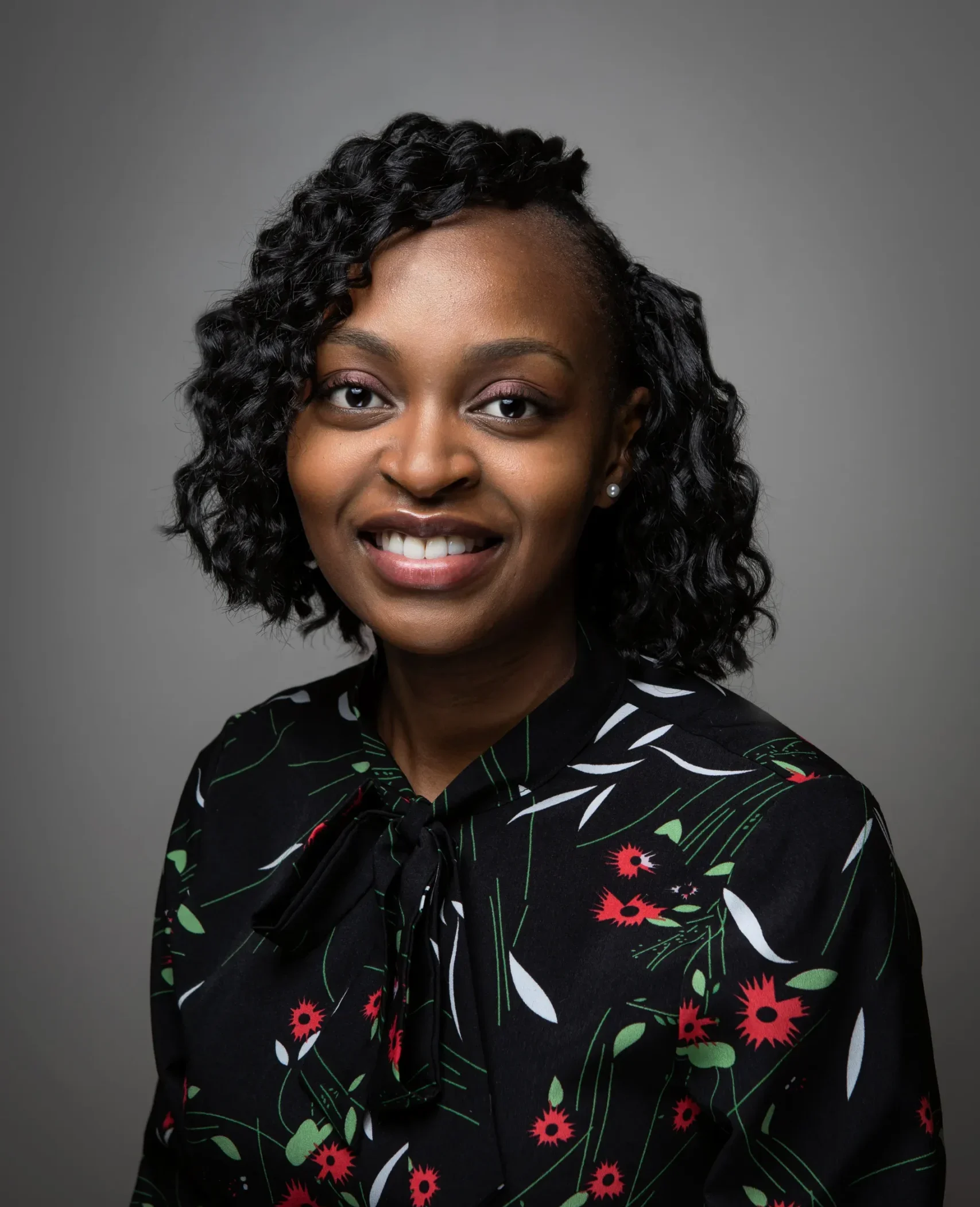 Scolastica Njoroge with curly dark hair smiles at the camera. She is wearing a black blouse with a floral pattern in red, green, and white, and is posed against a plain gray background. -Kaki Foundation