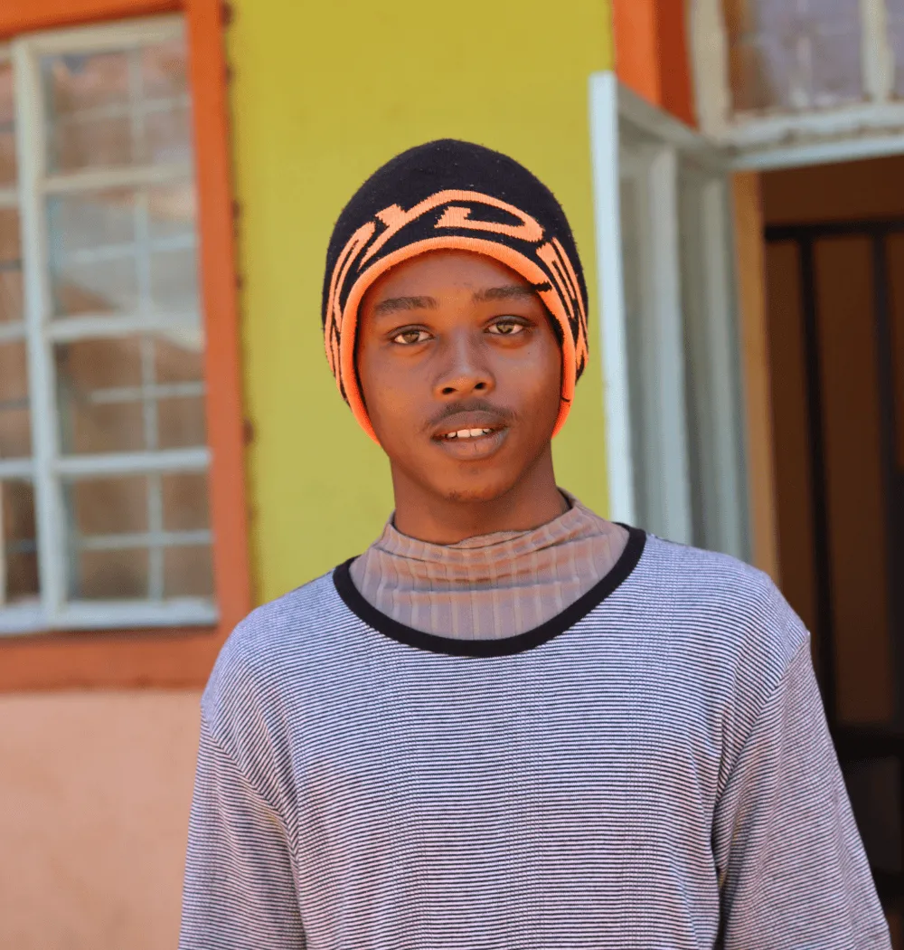 A young person wearing a striped shirt and a black and orange beanie stands outside in front of a yellow and orange building with an open door and window. -Kaki Foundation