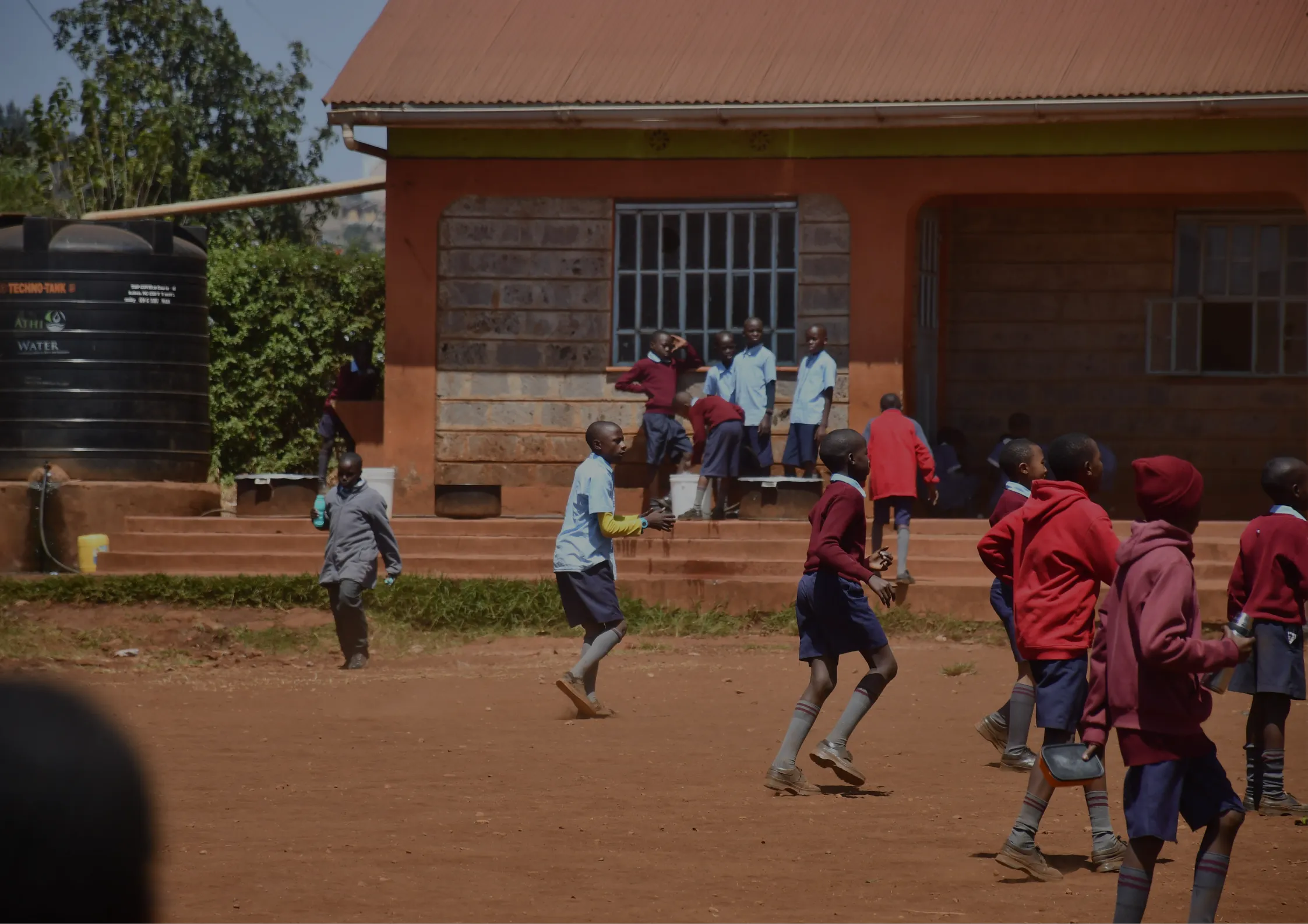 Children in school uniforms play outside on a dirt yard in front of a brick school building. Some children run and others stand near the steps, enjoying their recess under a sunny sky. -Kaki Foundation