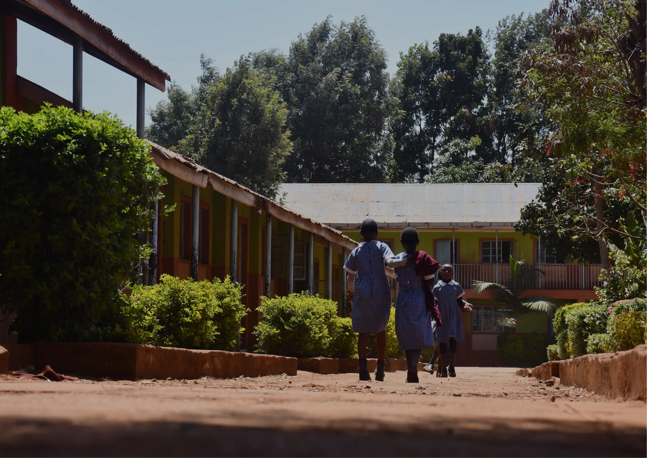 Children in school uniforms walk along a dirt pathway between two classroom buildings surrounded by greenery, with trees and a bright sky in the background. -Kaki Foundation
