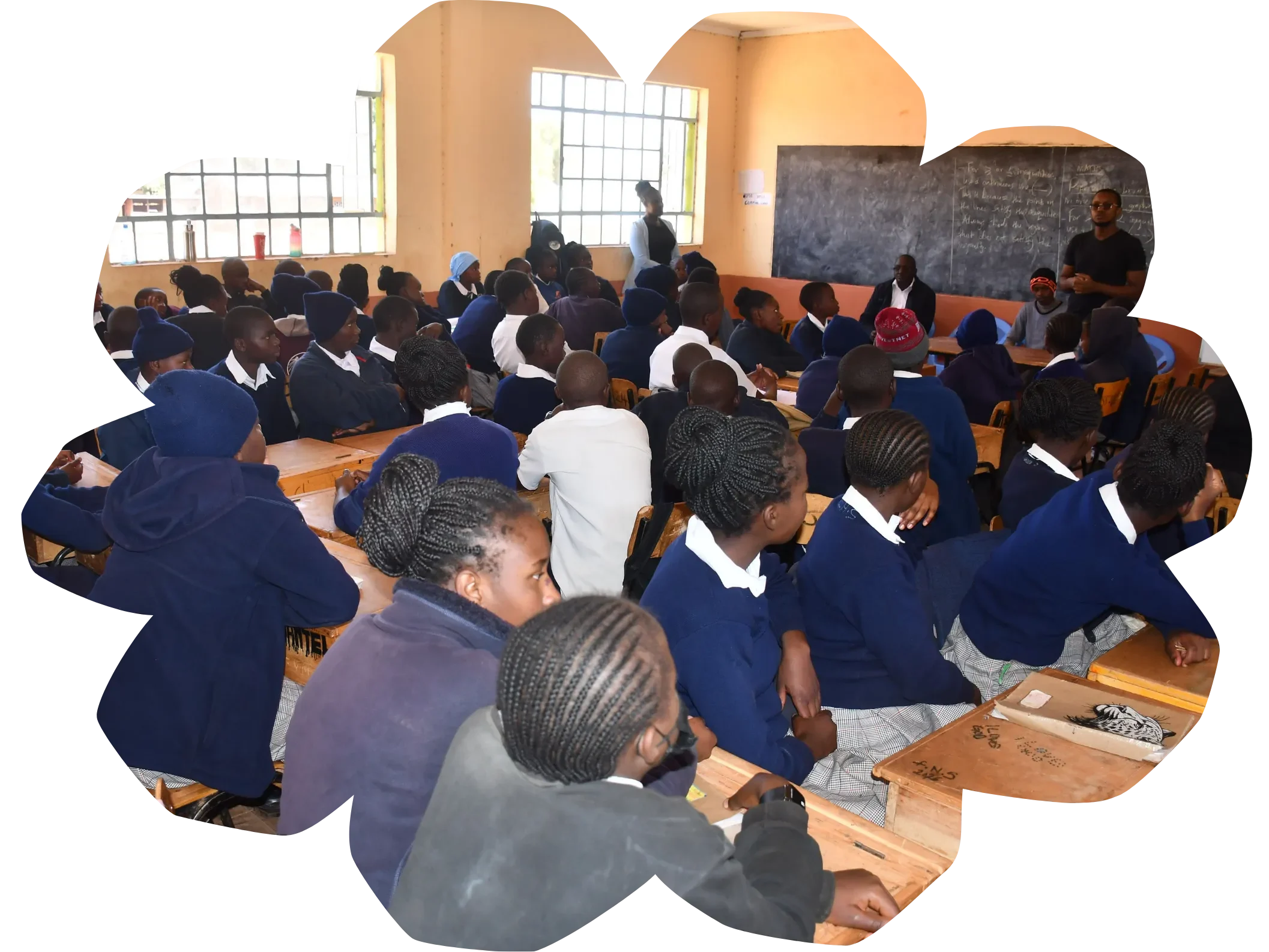 A classroom filled with students in blue uniforms sitting at desks, attentively facing a teacher at the front near a chalkboard, in a brightly lit room with large windows. -Kaki Foundation