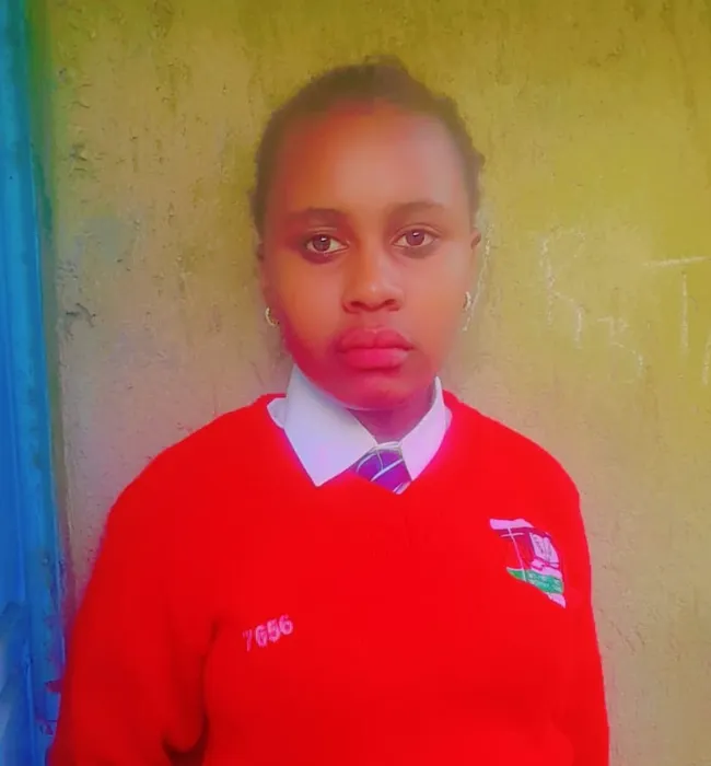 A student with short hair, wearing a red jacket with cartoon characters, stands against a plain blue background, looking directly at the camera with a neutral expression—one of the inspiring Kaki Foundation Kenya scholarship success stories. -Kaki Foundation