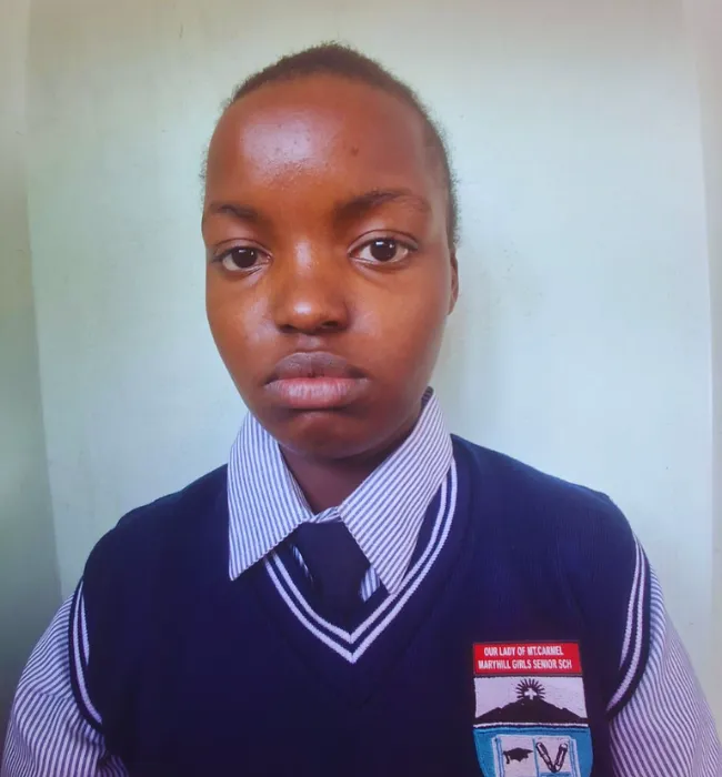 A student in a navy blue school uniform stands in front of a leafy green background, representing Kaki Foundation Kenya scholarship success stories. The uniform has a striped collar and a badge; the student looks directly at the camera with a neutral expression. -Kaki Foundation