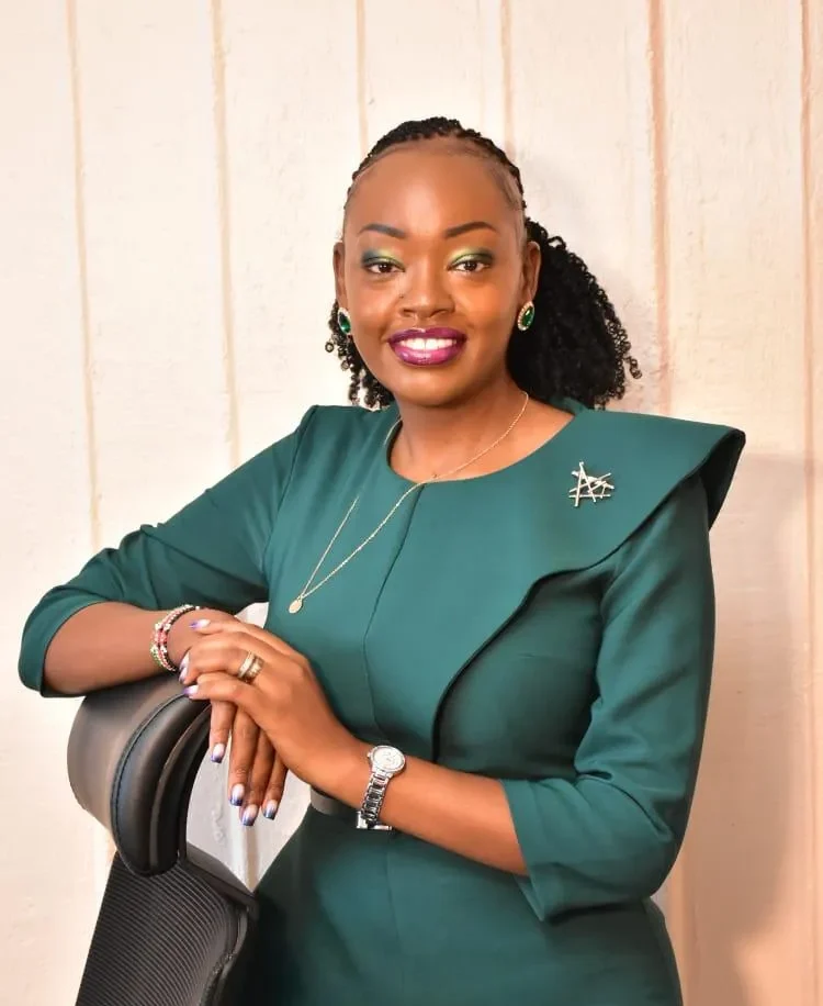 A woman with braided hair, wearing a green dress and jewelry, smiles while leaning on a black chair against a light-colored wall. -Kaki Foundation